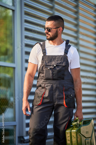 Strong gas station worker holding canister full of fuel. Male operator caring heavy cistern with gasoline. Confident man in sunglasses and overalls with canister looking away.