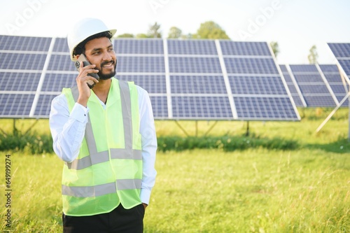 young male technician worker wearing green vests and helmet talking on phone about the maintenance and installation of the solar panels standing in field, technology in agriculture © Serhii