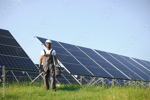 Competent energy engineer in grey overalls and white helmet checking solar panels while walking on field. African american man carrying clipboard and container with instruments