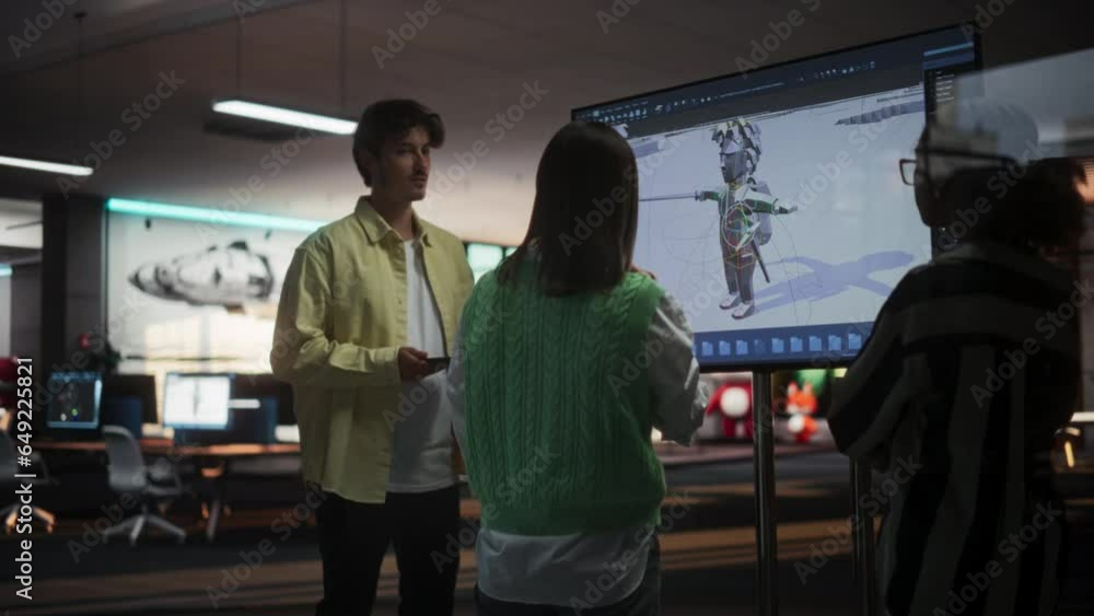 Multiethnic Male And Female Colleagues Standing Next To TV Set With 3D Model Of Video Game Character In Game Development Studio Office. Diverse Game Designers Discussing New RPG Game Character. Multiethnic Male And Female Colleagues Standing Next To TV Set With 3D Model Of Video Game Character In Game Development Studio Office. Diverse Game Designers Discussing New RPG Game Character.