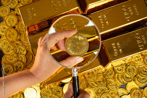 a hand with a magnifying glass checks the Bitcoin on a pile of gold coins and gold bars