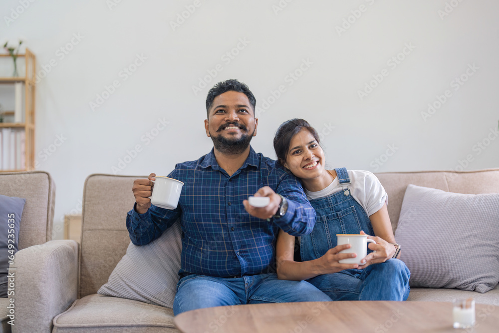 Indian couple watch tv on the couch at their living room, husband and wife spending quality time together pleasure smile. man holding remote control at hand. Concept weekend