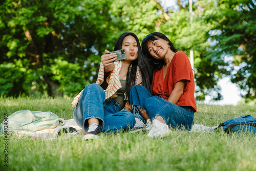 Two asian women taking selfie while sitting in green park