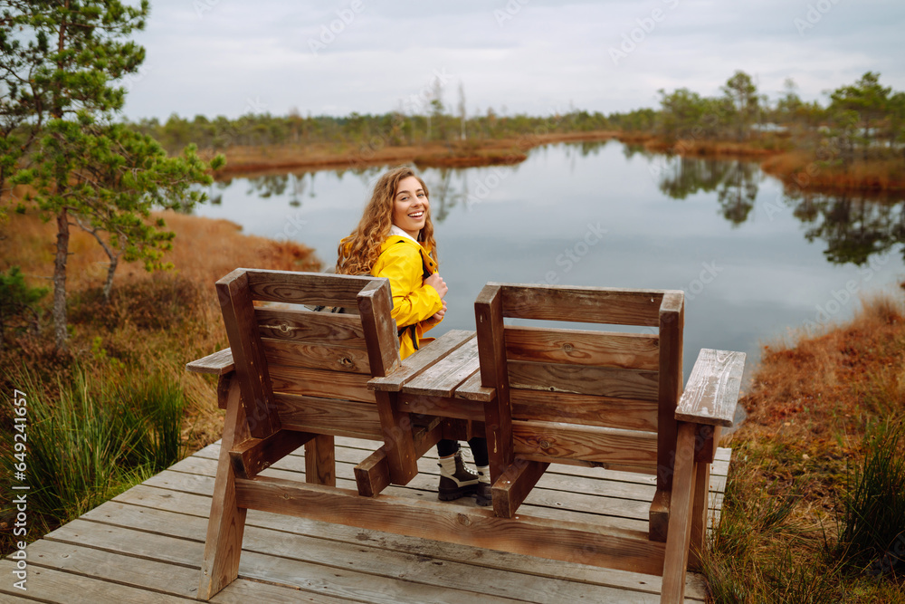 A hiker female wearing a yellow coat and backpack walks along a scenic nature trail with a wooden boardwalk overlooking wetlands. Travel and exploration.