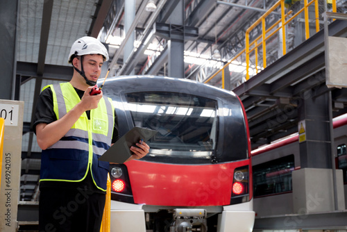 Young caucasian engineer man or worker hold clipboard for looking and checking electric train for planning maintenance and using radio for command in station, inspector check service transport.