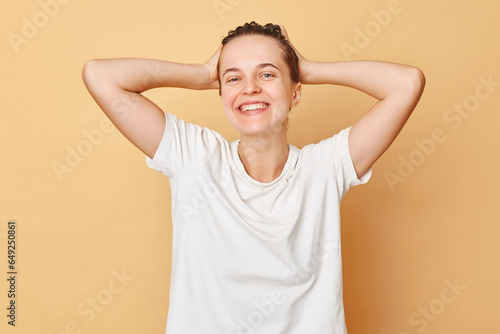 Healthy hair. Wellbeing wash. Pampering routine. Satisfied smiling woman wearing white T-shirt washes hair standing with shampoo foam on her head isolated over beige background