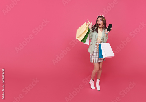 Trendy beautiful young Asian teen woman carrying colorful paper bags shopping online with mobile phone and hand pointing finger isolated on pink banner background with copy space