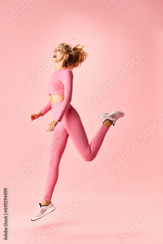 Portrait of happy, joyful slim woman running to fintess traing, gym dressed in sport cloth isolated over pink background