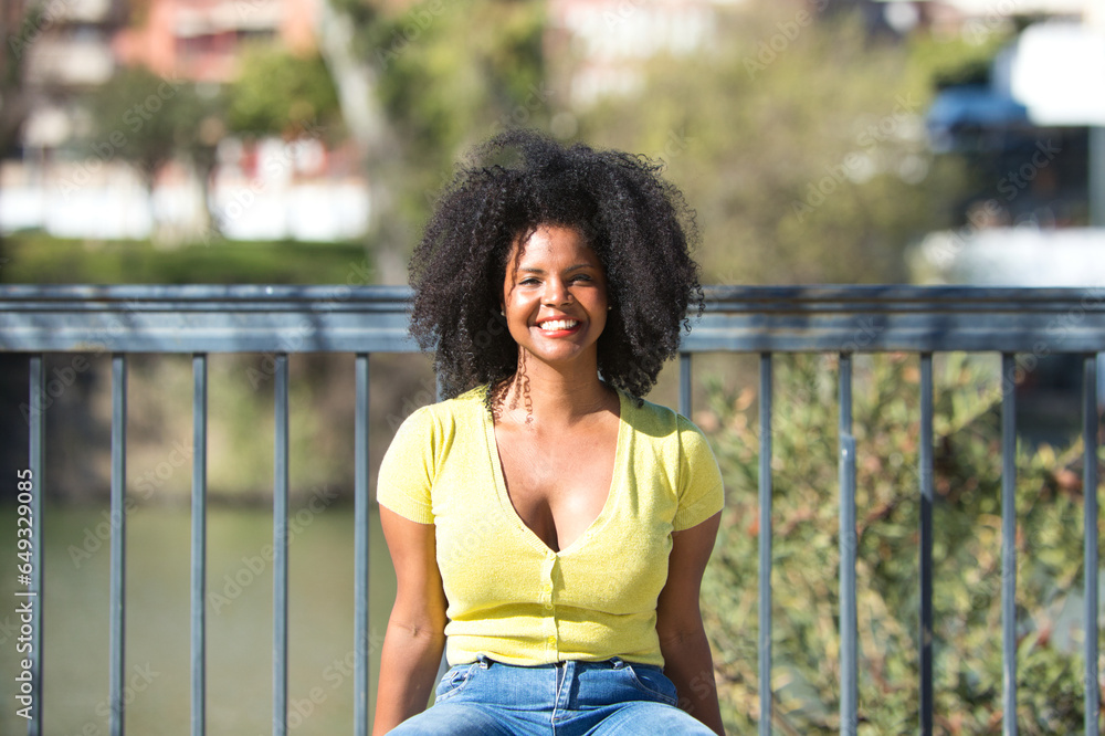 Young and beautiful black woman with afro hair dressed in jeans and yellow shirt sitting on a wooden bench, the woman is happy. In the background the river guadalquivir of seville in spain. Young and beautiful black woman with afro hair dressed in jeans and yellow shirt sitting on a wooden bench, the woman is happy. In the background the river guadalquivir of seville in spain.