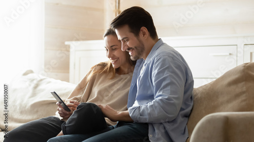 Happy man and woman using phone together, sitting on couch, watching video in social network or chatting, browsing apps, smiling young couple shopping online, spending leisure time on cozy sofa