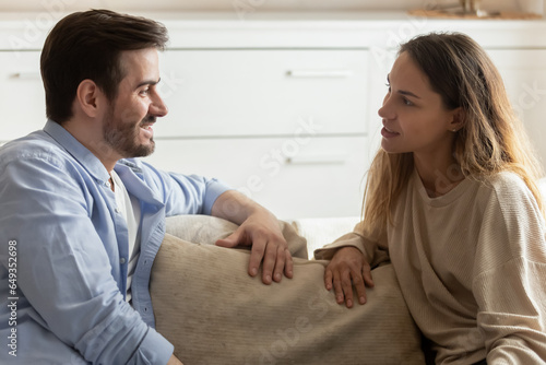 Happy young man and woman chatting, sitting on couch in living room, wife and husband talking, enjoying leisure time at home together, friends having pleasant conversation, sharing news