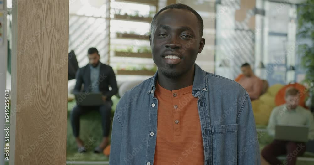 Slow motion portrait of young African American man standing in modern comfortable coworking space smiling looking at camera. Job and workplace concept. Slow motion portrait of young African American man standing in modern comfortable coworking space smiling looking at camera. Job and workplace concept.