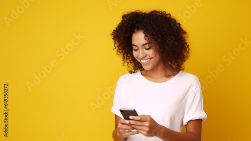Latin woman wearing a white t-shirt and holding mobile phone looking at smartphone isolated on yellow background