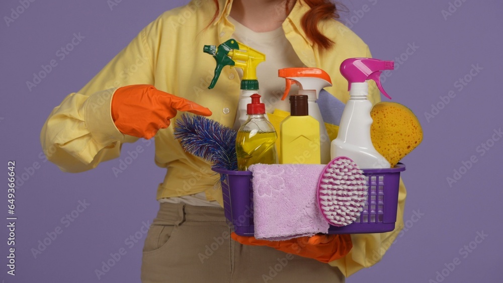 Closeup shot of woman in rubber gloves holding basket with cleansers and sponges, pointing at it. Isolated on purple background.