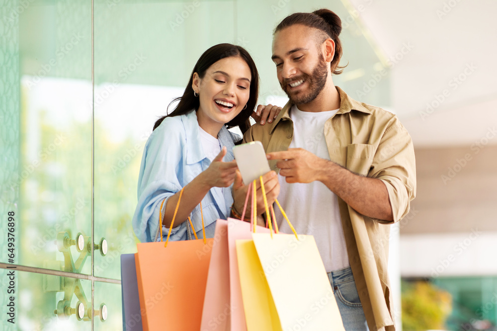 Cheerful surprised young european couple with bags enjoy shopping and spare time, looking at smartphone
