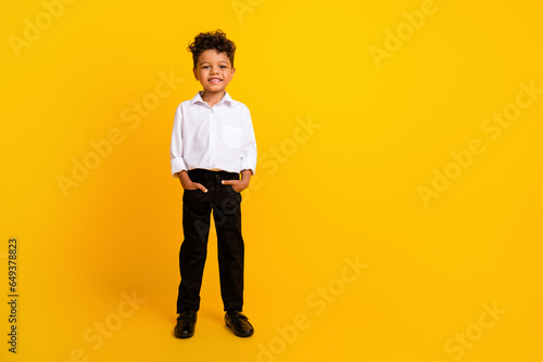 Portrait of pleasant funny schoolboy with wavy hair wear stylish shirt hold palms in pockets standing isolated on yellow color background