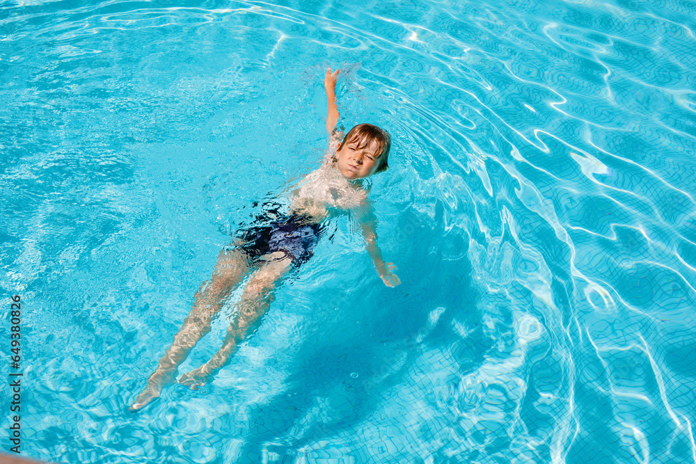 Portrait of happy little kid boy in the pool and having fun on family vacations in a hotel resort. Healthy child playing in water, swimming and splashing.
