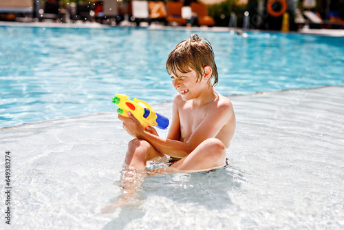 Happy little kid boy jumping in the pool and having fun on family vacations in a hotel resort. Healthy child playing in water with a water gun. Laughing running child splashing