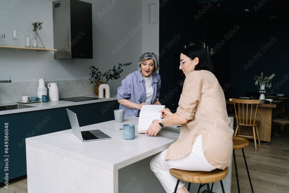 Naklejka premium Surprised elderly woman watching video on computer while daughter working in kitchen