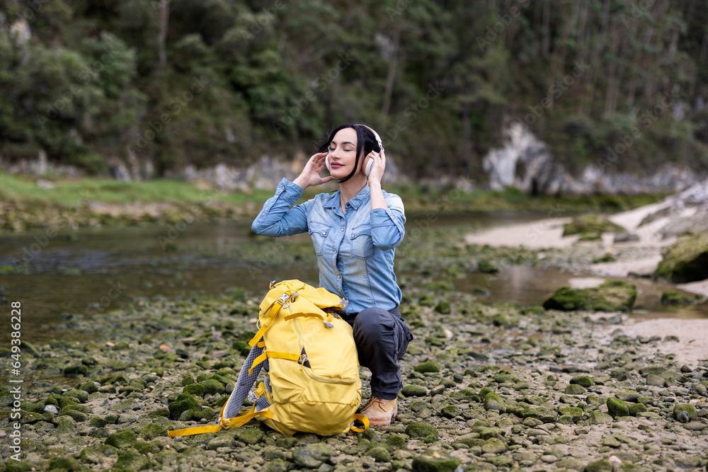 Young adult woman in the middle of the forest next to a river, listening to music with her headphones
