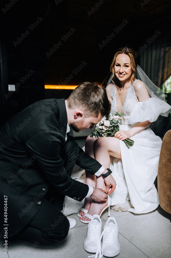 A young stylish groom helps a bride in a white dress to change shoes, put on shoes indoors. Wedding photography, portrait.