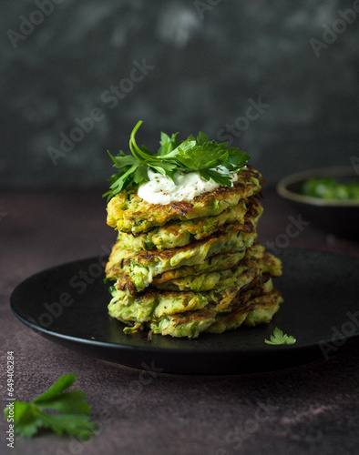 A stack of zucchini pancakes with sour cream and parsley on a black ceramic plate, dark style.