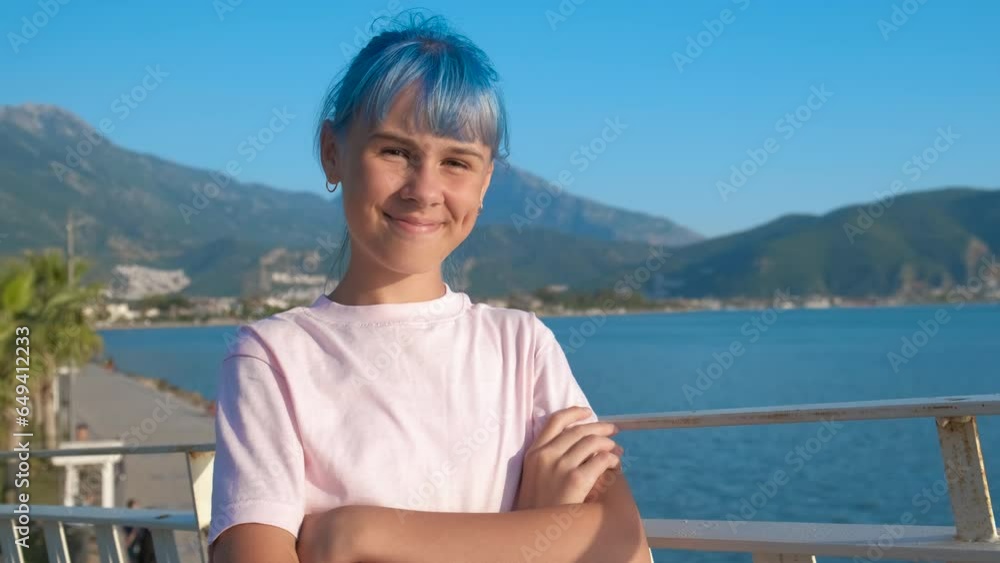 Stylish girl stand against blue bay. A stylish blue haired teen stand on the river bank against turquoise mountains in the sun light.
