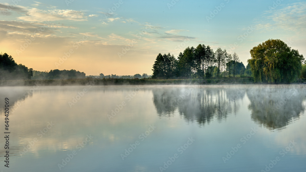A picturesque sunrise over a foggy lake with trees on the shore reflected in the water. Morning of the end of summer, beginning of autumn.