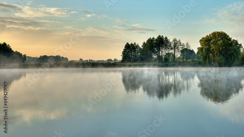 A picturesque sunrise over a foggy lake with trees on the shore reflected in the water. Morning of the end of summer, beginning of autumn.