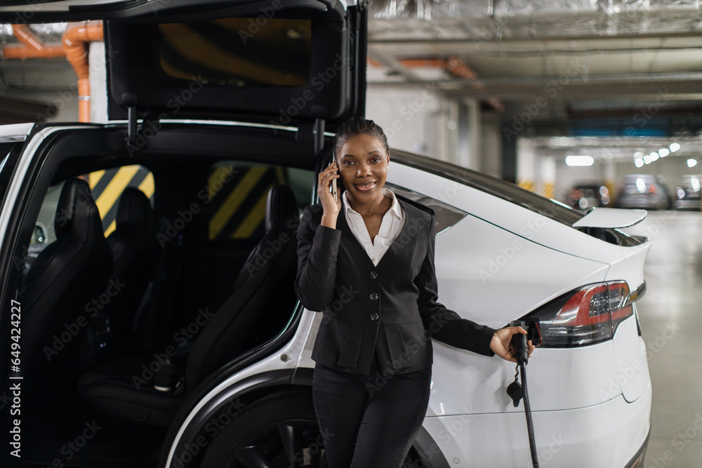 Φωτογραφία African female listening to phone caller whilt filling up electric motorcar from EV charger in basement garage