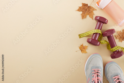 Autumn style and fitness harmony. Top view photo of stylish sneakers, measuring tape, dumbbells, plastic water bottle, fallen maple leaves on pastel brown background with promo spot