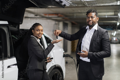 Foto Portrait of african man and woman with smartphones posing near EV in charging session during rental procedure