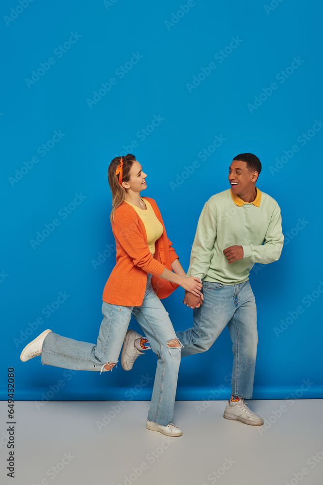 Naklejka premium happy multicultural couple in casual attire holding hands and running on blue background, smile