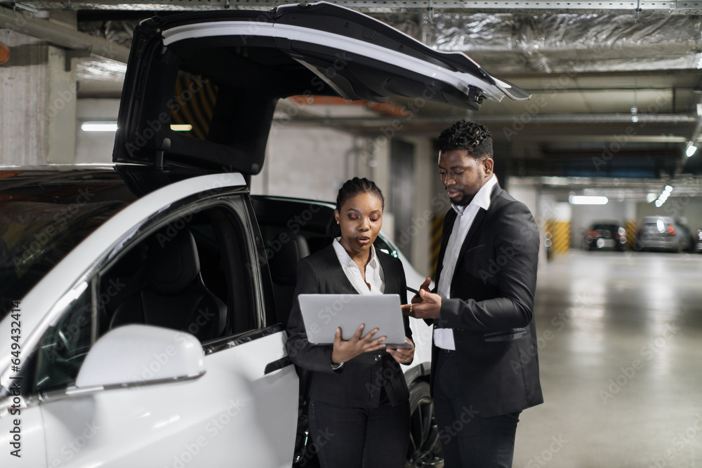 African working mates in formal outfits looking at laptop screen in underground garage with e-car. Commercial partners inspecting financial report via modern technologies in bottom-level parking. African working mates in formal outfits looking at laptop screen in underground garage with e-car. Commercial partners inspecting financial report via modern technologies in bottom-level parking.