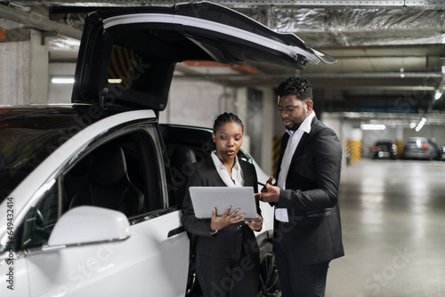 African working mates in formal outfits looking at laptop screen in underground garage with e-car. Commercial partners inspecting financial report via modern technologies in bottom-level parking.