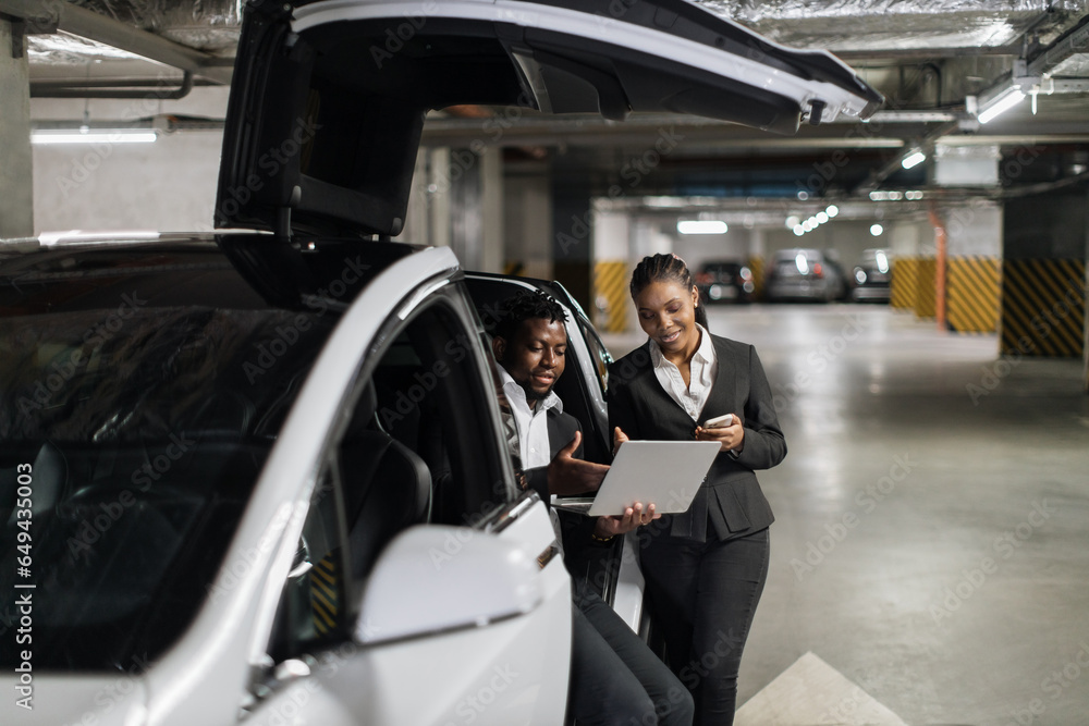 African american man and woman operating laptop while staying inside EV in bottom-level garage. Professional partners in formal attire managing financial assets beyond office walls.