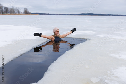 Winter swimming. Woman in frozen lake ice hole. Swimmers wellness in icy water. How to swim in cold water. Beautiful young female in zen meditation. Gray hat and gloves swimming clothes. Nature lake