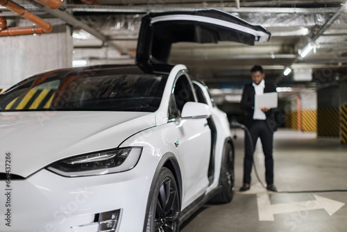 Focus on electric motorcar with door open vertically being refueled via electric port by male owner with gadget in garage. Effective entrepreneur answering invoices via wireless connection indoors.