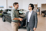 Portrait of professional salesman in business suit shaking happy client hand and talking. Stylish male car dealer serving smiling customer in showroom. Car business, trading, dealership.