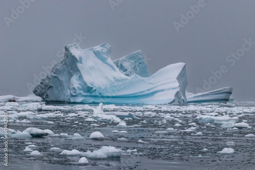 Iceberg with blue and white ice near the antarctic peninsula. Snowy gray sky in background; calm water in foreground covered with ice flow. 
