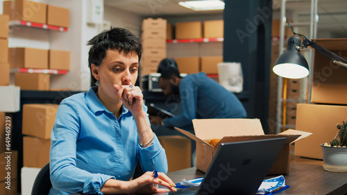 Woman employee analyzing goods from warehouse shelves, looking at merchandise in cardboard boxes before shipping goods order. Young adult working in storage space with supply chain.