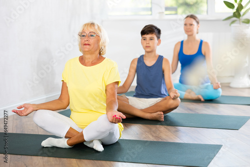 Grandmother, mom and teen son do half lotus position of Ardha Padmasana Sidhasana with hands clasped in mudra of knowledge in gym. Family three generations keep fit with exercise. Healthy lifestyle
