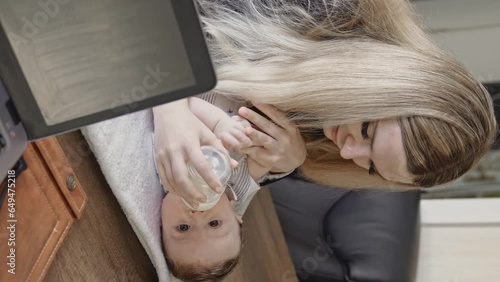 Business woman feeds baby from a bottle in office on desktop, concept of working after birth of child, vertical video photo