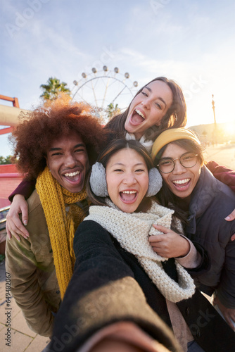 Vertical excited smiling group multiracial friends taking funny selfie looking at camera standing together outdoor. Multi-ethnic young people in amusement park having fun in sunny winter holiday.  photo