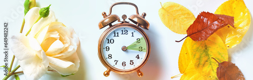 Vintage alarm clock showing the hour between daylight saving time in summer and fall back symbolized by a rose on one side and autumn leaves on the other, panoramic format