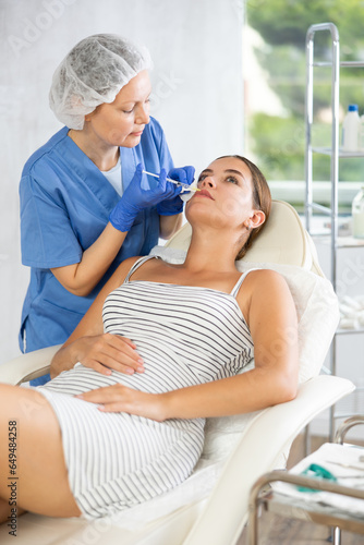 Adult female cosmetologist in uniform makes injection in lips of young woman patient in medical room