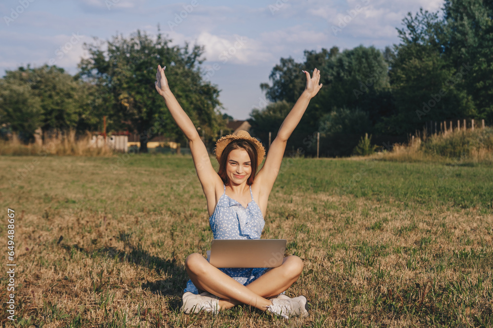 Beautiful caucasian girl with long hair in the blue dress and summer hat working on the laptop, remote work at the countryside. Banner, place for text concept.