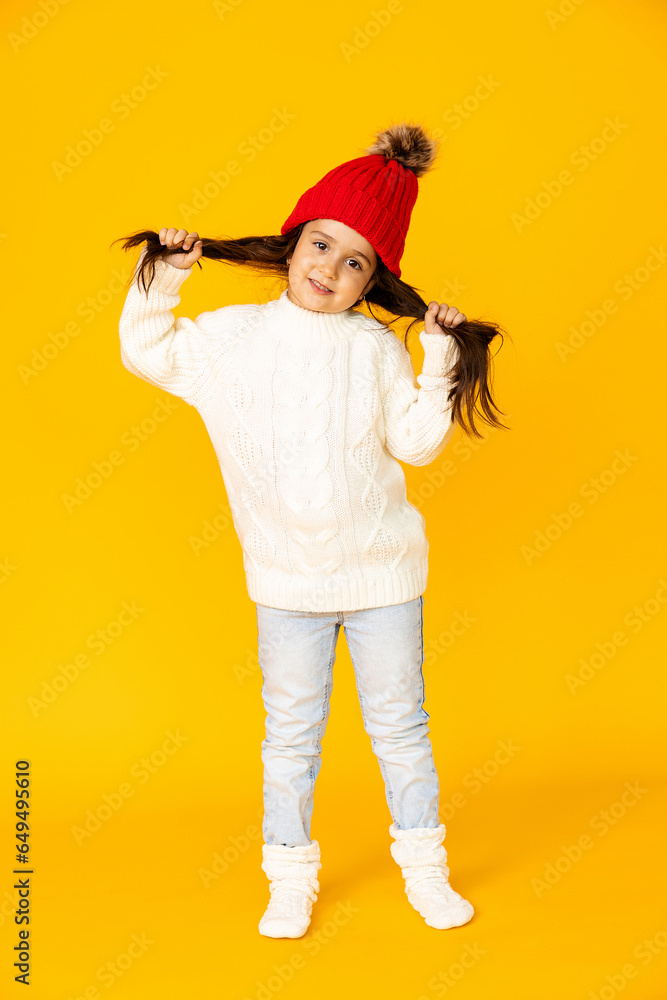 Funny child girl in a white sweatshirt and red knitted hat with pompom on a yellow background. The concept of children's emotions, joy and delight. Advertising clothing and sales. Ski resort vacation Funny child girl in a white sweatshirt and red knitted hat with pompom on a yellow background. The concept of children's emotions, joy and delight. Advertising clothing and sales. Ski resort vacation