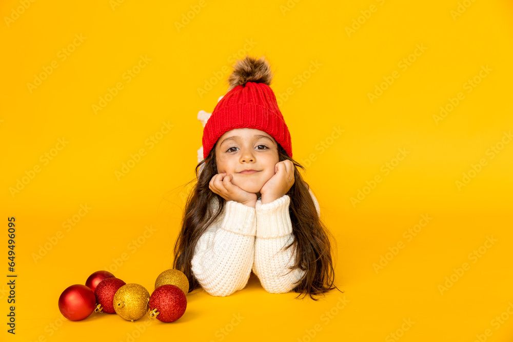 Funny child girl in a white sweatshirt and red knitted hat with pompom on a yellow background. The concept of children's emotions, joy and delight. Advertising clothing and sales. Ski resort vacation