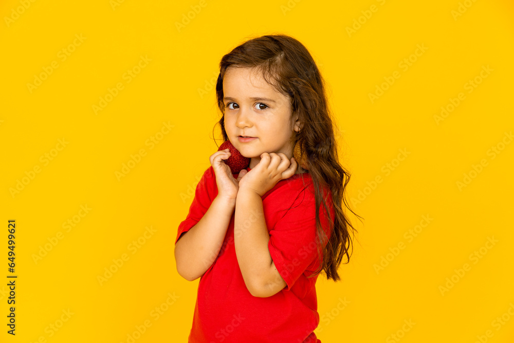 A cute little girl in a red t-shirt holding Christmas tree toys. yellow background, space for text A cute little girl in a red t-shirt holding Christmas tree toys. yellow background, space for text